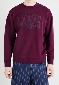Sudadera marrón con cuello redondo y logo texturizado de Levi's en la parte frontal. Mangas largas y un corte holgado. Combinada con pantalones azul a rayas.