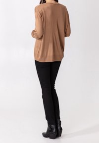 Ruskea neulottu pitkähihainen rento neule, jossa takasauman yksityiskohta; yhdistetty mustiin kapealahkeisiin farkkuihin ja mustiin nilkkureihin.