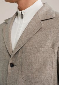 Rjava jakna iz mešane volne z razrezanim ovratnikom, enojnim črnim gumbom in žepom na prsih. Nosijo jo čez belo zip-up jopo.