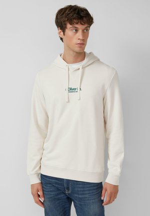 Young man wearing off-white hoodie with green text and blue jeans, looking to the side against a plain light gray background.