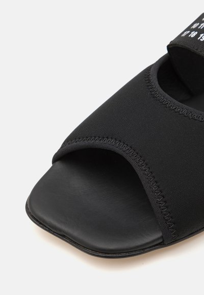 Sorte sklippesandal med en myk, elastisk tekstiloverdel og åpen tå-design, samt en teksturert svart såle. Utsmykket med hvit tekst.