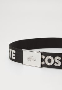 Ceinture tissée noire avec un texte blanc contrastant et un fermoir en métal argenté portant un logo de crocodile. Surface lisse et conception robuste.