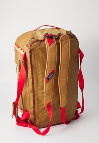 Rectangular tan backpack with red straps, padded shoulder straps, a top handle, and a Patagonia logo on one strap.