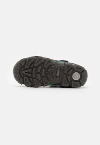 Semelle de chaussure en caoutchouc noir avec des motifs de grip texturés, des lignes d'accent vertes et un logo circulaire. Présente un design durable et robuste pour une meilleure traction.