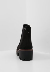 Černé semišové kotníkové boty s masivní černou podrážkou a hnědým akcentem, vybavené elastickými postranními panely a poutkem na zadní straně.