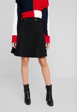 Woman wearing a black A-line skirt with button details, a color-block red, white, and navy sweater, and black ankle boots standing on a plain background.