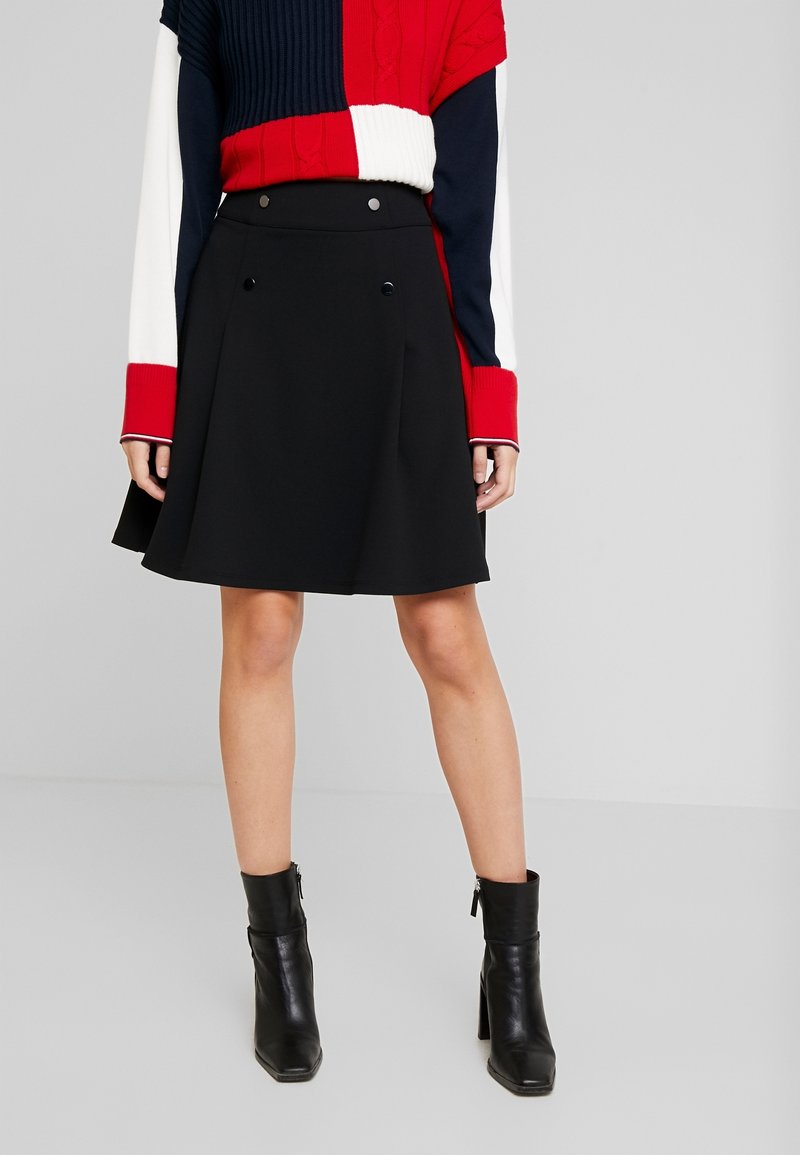 Femme portant une jupe noire trapèze avec des détails de boutons, un pull color-block rouge, blanc et bleu marine, et des bottines noires, debout sur un fond uni.