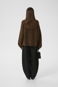 Kvinne med langt brunt hår iført en løs brun genser, vide svarte bukser, og som holder en svart håndveske, står mot en ensfarget bakgrunn.