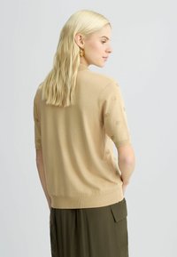 Beige gebreid trui met korte mouwen, met een gestructureerde afwerking en knoopaccenten aan de zijkant, weergegeven op een model van achteren.