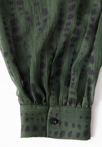 Blusa verde a pieghe con macchie nere, caratterizzata da un polsino con bottoni e una texture leggera e trasparente. Cuciture decorative visibili all'orlo.
