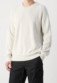 Krémový svetr s texturou a ribbovaným designem, kulatým výstřihem a subtilní nášivkou s logem. Rukávy jsou mírně nadměrné.