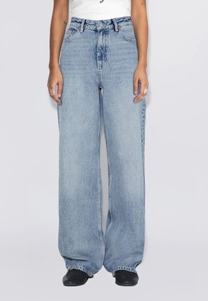 Persoon die lichtblauwe wijde jeans, een wit topje, zwarte schoenen en zilveren ringen aan beide handen draagt, staand tegen een effen achtergrond.