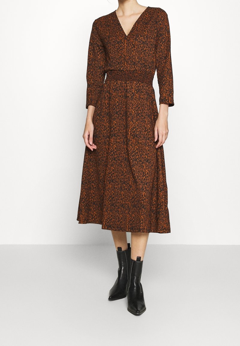 Femme portant une robe midi marron et orange à motifs avec un col en V, des manches longues et des bottines noires sur un fond blanc uni.