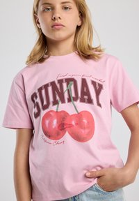 Camiseta de algodón rosa con un gráfico de dos cerezas, un gran texto "SUNDAY" y la frase "California Cherry" en una fuente cursiva.