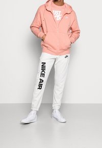 Rosa zip-up hoodie med ficka fram, vit t-shirt med "Just Do It" grafik, vita sweatpants med svart "NIKE AIR" tryck, och vita sneakers.