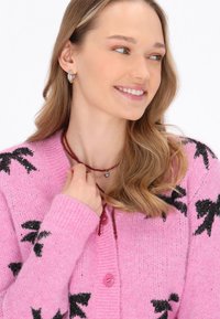 Cárdigan de punto rosa con patrones de lazo negro, con un escote redondo y botones rosas. Collar de cuentas rojas con un colgante en forma de corazón.