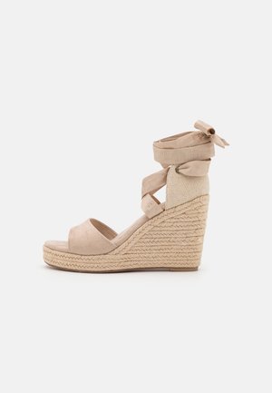 Beige kile-sandal med fletplatform i jute, åben tå og stofremme omkring anklen, bundet i en sløjfe bagpå.