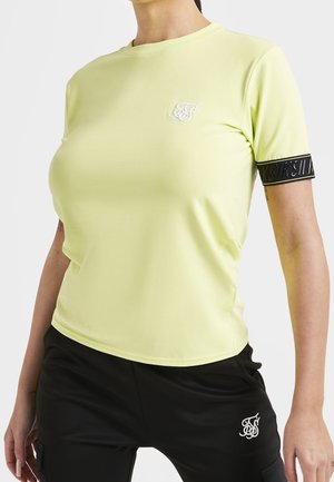 Mujer que lleva una camiseta ajustada de manga corta amarillo claro con detalles de logo en negro, combinada con pantalones negros con logo blanco.