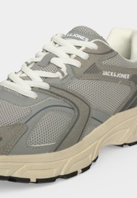 Γκρι και μπεζ sneaker Jack & Jones με λευκά κορδόνια, ύφασμα από διχτυωτό υλικό, σουέντ λεπτομέρειες και υφή στην σόλα, εμφανίζεται από πλάγια κοντινή γωνία.