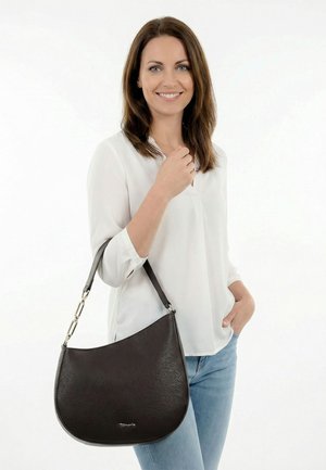 Femme portant un chemisier blanc et un jean, souriant en tenant un sac à bandoulière en cuir noir avec des détails métalliques.