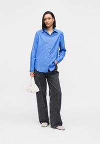 Blau gestreiftes Hemd mit Knöpfen, graue Hosen mit weitem Bein, beigefarbene Schuhe und eine weiße Handtasche mit Plisseefalten und einer strukturierten Oberfläche.