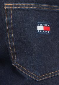 Τσέπη από σκούρο μπλε τζιν με πορτοκαλί ραφές και μια μικρή ορθογώνια ετικέτα με τ ο λογότυπο Tommy Jeans σε λευκό, κόκκινο και ναυτικό μπλε χρώμα.