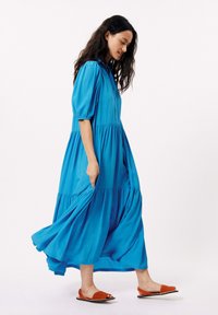 Blauwe katoenen maxi-jurk met pofmouwen, knoopsluiting aan de voorkant en gelaagde rok. Gepaard met oranje platte sandalen, wat een levendig contrast creëert.