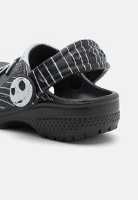 Black clogs with a Jack Skellington design, featuring white stripes and a round, smiling skull accent on the side, made from a lightweight material.