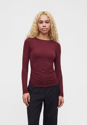 Femme aux longs cheveux blonds bouclés portant un haut bordeaux ajusté à manches longues et un pantalon noir, debout devant un fond clair uni.