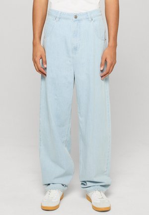 Personne portant un pantalon large à rayures bleu clair avec poches avant et des baskets blanches, debout devant un fond uni.