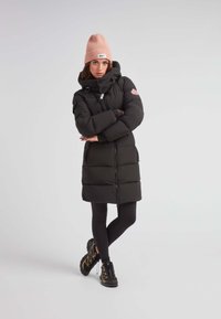 Abrigo acolchado negro con capucha, que presenta un acento rosa en la manga y bolsillos con cremallera. Combinado con leggings negros y zapatillas negras.