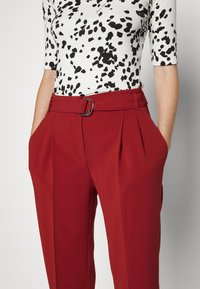 Red pleated trousers with a wide waistband, featuring a metal buckle. Paired with a black and white spotted patterned top.