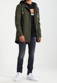 Parka à capuche vert olive avec fermeture éclair et boutons-pression, deux poches frontales, portée sur un sweat-shirt noir, associée à un jean foncé et des baskets blanches.