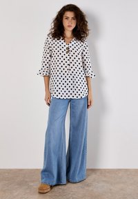 Weiße Bluse mit Polka-Dots und dreiviertel Ärmeln, V-Ausschnitt und drei Knöpfen vorne, kombiniert mit weit geschnittenen blauen Jeans.