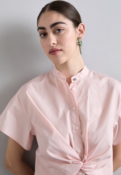 Blusa a maniche corte di colore rosa chiaro con chiusura a bottoni e dettaglio a nodo intrecciato in vita. Tessuto liscia e design del colletto strutturato.