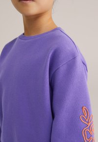 Hanorac mov cu guler rotund, umeri căzuți și design brodat portocaliu pe mânecă stângă. Textură moale a materialului.