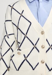 Cárdigan de punto en crema con patrón diagonal en azul marino. Presenta botones redondos de plata y un bolsillo en la parte frontal. Textura suave y ajuste entallado.