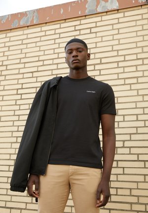 Black cotton t-shirt with short sleeves and white logo on the chest, paired with khaki pants and a black lightweight jacket, against a brick wall.
