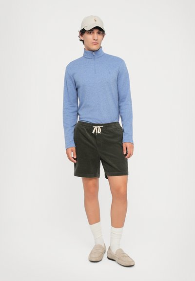 Camisola azul com fecho éclair de mangas longas, calções de veludo cotelê verde-oliva, meias brancas e mocassins bege; apresenta um logótipo no boné e cordão nos calções.