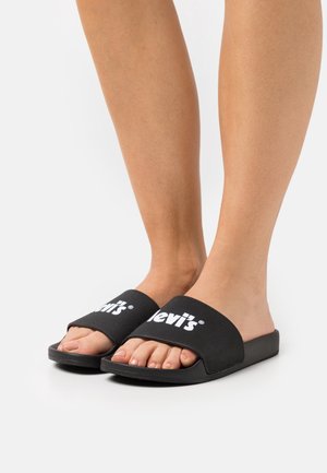 Persoon met zwarte Levi's schuifsandalen met witte logo, staand op een witte achtergrond, met blote voeten en benen.
