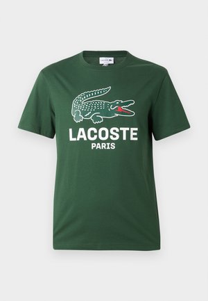 Zelena majica s kratkimi rokavi z veliko belo in rdečo grafično podobo krokodila nad napisom "LACOSTE PARIS" na sprednji strani.