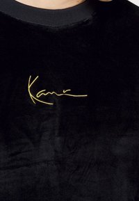 Černá sametová mikina s kulatým výstřihem, na levé straně hrudi s žlutým vyšitým podpisem. Měkká textura.