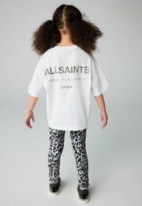 Bijela oversized majica s natpisom "ALLSAINTS" u teksturiranom crnom fontu, u kombinaciji s leggings hlačama u crno-bijelom leopard uzorku i crnim cipelama.