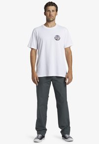 Hvid bomulds t-shirt med rund halsudskæring, prydet med et cirkulært grafisk motiv på brystet, sat sammen med mørkegrå bukser og sorte sneakers.
