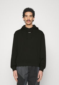 Sudadera negra con capucha y cordones, que presenta un logo blanco "HUGO" en el pecho. Tejido suave, corte relajado y puños acanalados.
