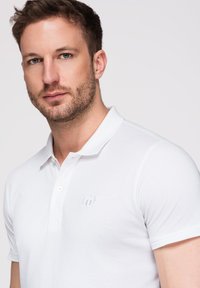 Homme aux cheveux bruns courts et à la barbe claire portant un polo blanc avec un petit logo brodé sur la poitrine.
