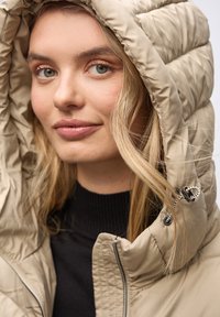 Beige pufferjakke med hætte, med en kviltet tekstur og lynlås lukning. Sort sweater synlig nedenunder, med hår der falder over skulderen.
