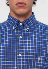 Blue and white checkered button-up shirt with a classic collar, featuring white buttons and a small embroidered logo on the left chest.