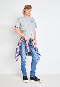 En ung person står klädd i en grå Levi's t-shirt, blå jeans, svarta högskor och en rutigt skjorta knuten runt midjan.