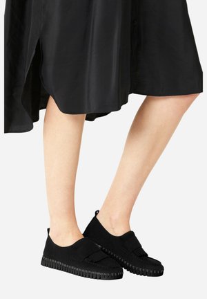 Zwarte suède schoenen met platte zolen, voorzien van een brede band over de voorkant en een gestructureerde rubberen buitenzool, gecombineerd met een zwarte jurk.